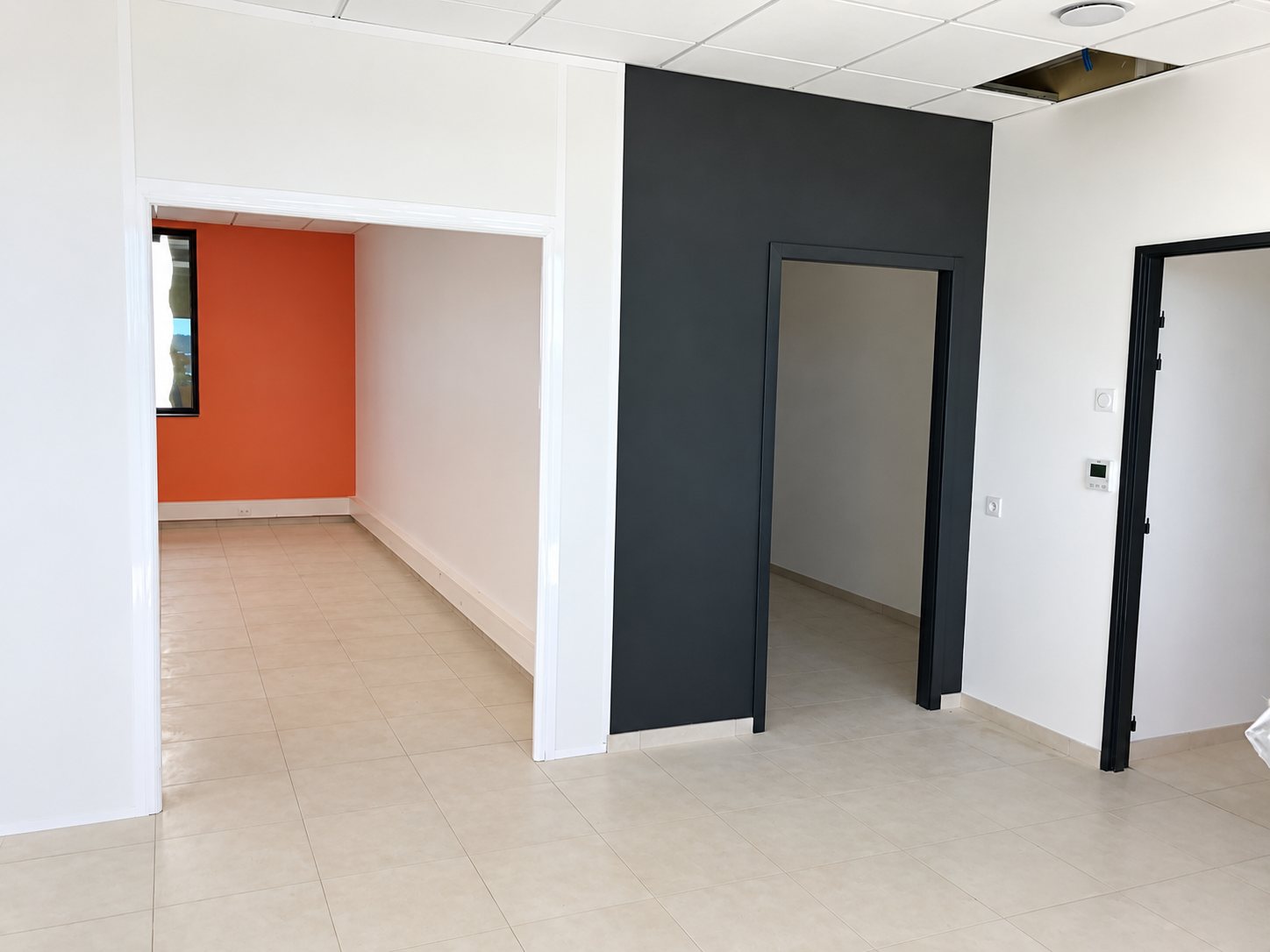 Pièce vide de bureaux récemment rénovés, avec cloisons blanches, un pan de mur gris anthracite, un mur orange au fond et un sol carrelé clair.