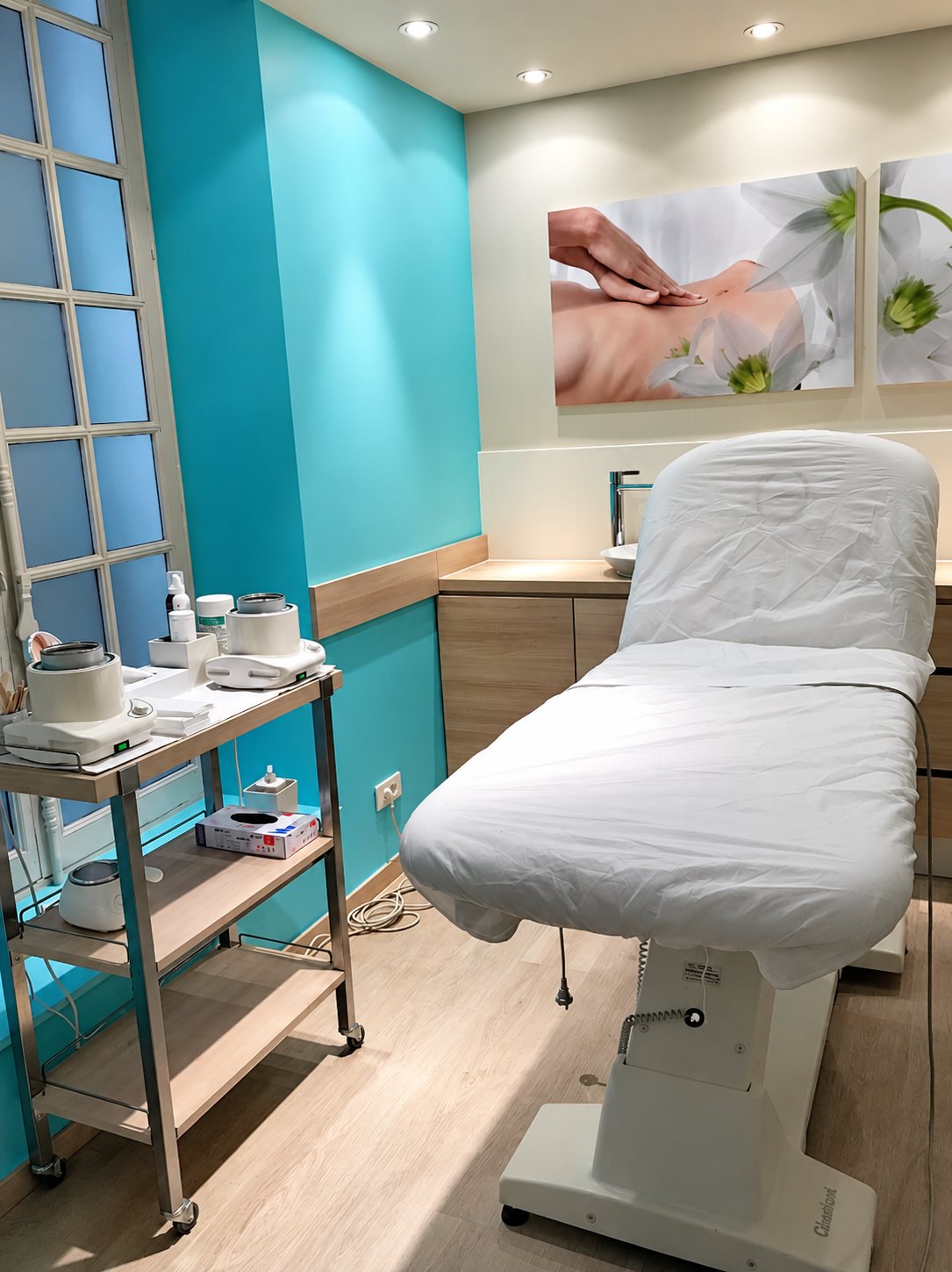 Cabine d'institut aux murs turquoise équipée d'une table de massage blanche, d'un chariot avec chauffe-cire et d'un grand tableau de fleurs.
