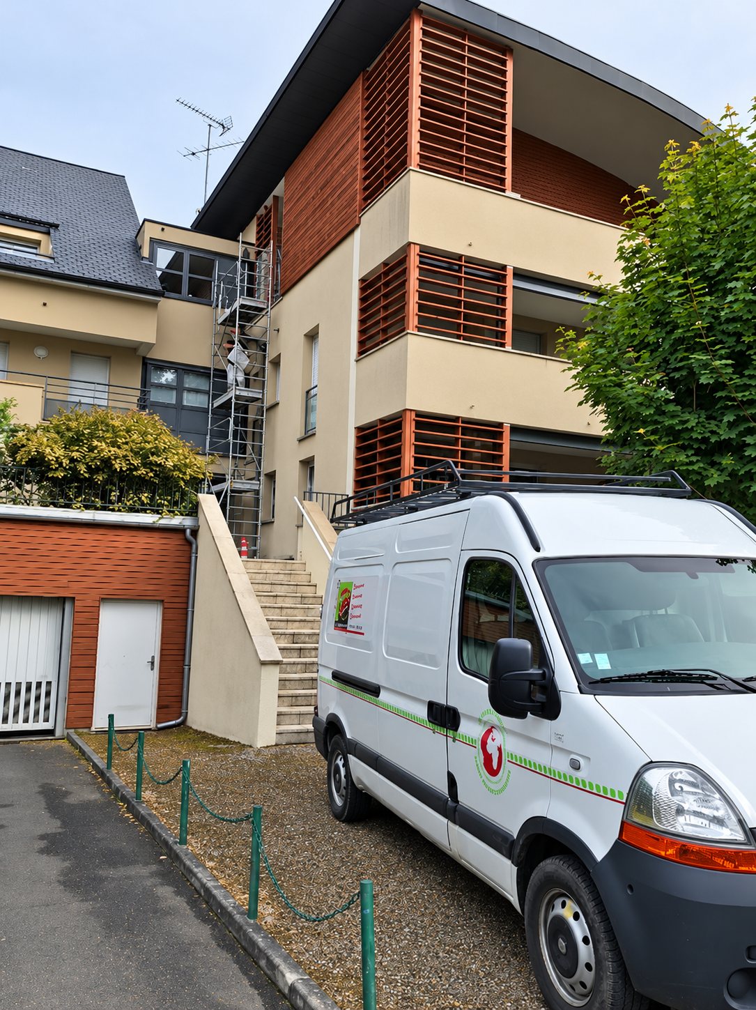Une camionnette blanche équipée d'une galerie est garée devant un immeuble résidentiel à persiennes en bois, où un échafaudage est dressé contre la façade.