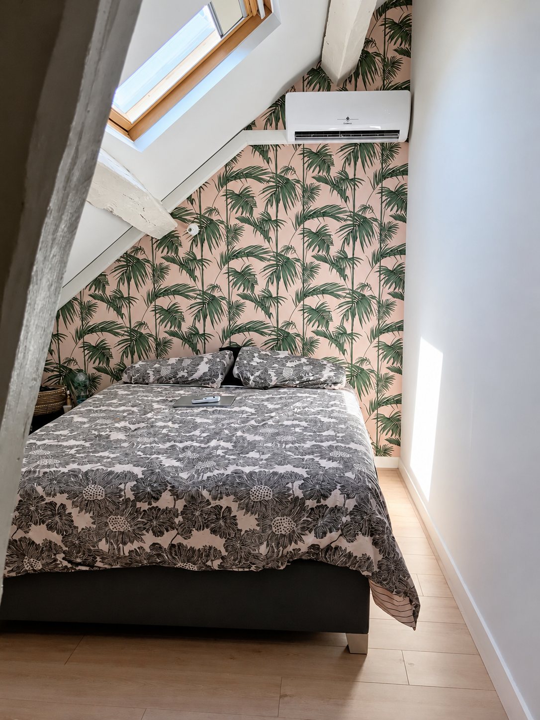 Chambre sous les combles avec un lit double, un papier peint à motif de palmiers verts sur fond rose, une fenêtre de toit et un climatiseur mural blanc.