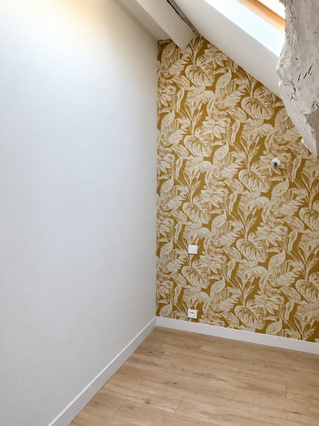 Pièce sous les combles fraîchement rénovée, avec un mur en papier peint à motifs de toucans et feuilles tropicales sur fond moutarde, un mur peint en gris clair et un sol en parquet stratifié.