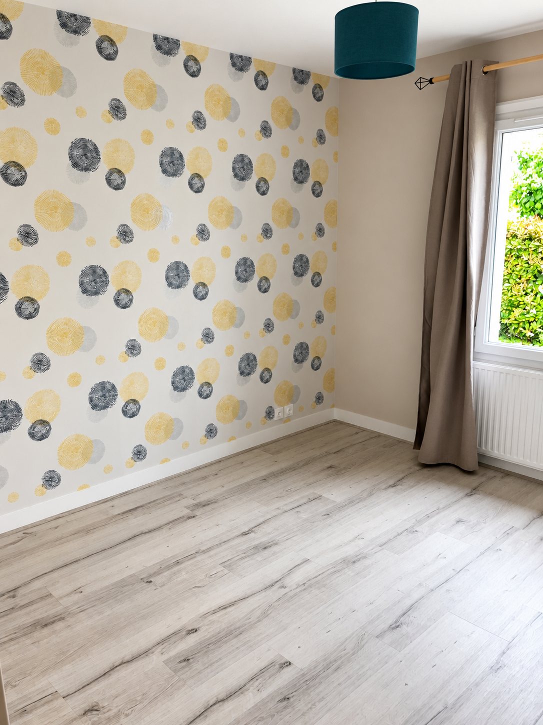 Chambre vide rénovée avec un mur en papier peint à motifs ronds jaunes et bleus, un sol stratifié gris clair, un rideau taupe et une fenêtre donnant sur un jardin.