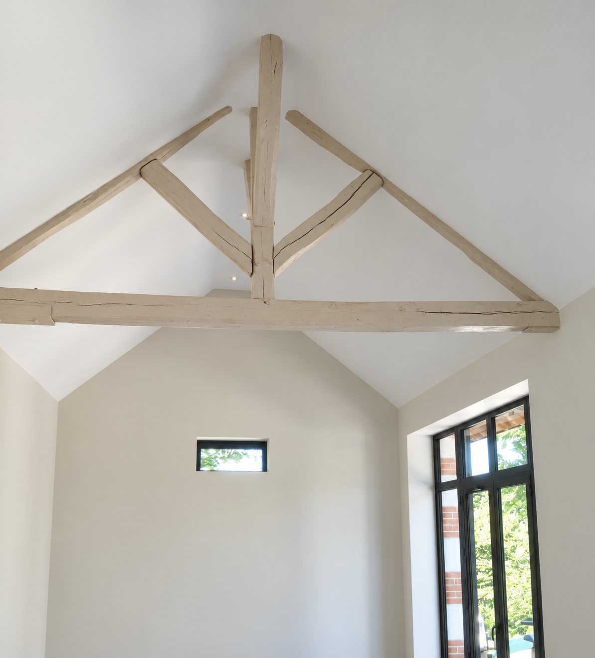 Une charpente apparente en bois peinte en blanc sous un haut plafond, avec une petite fenêtre et une grande porte-fenêtre donnant sur un jardin.