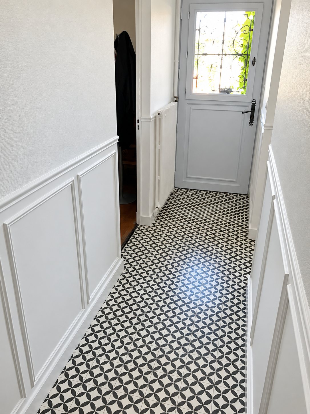 Un couloir étroit rénové avec un sol en carreaux de ciment à motifs noir et blanc, des soubassements blancs moulurés et une porte d'entrée vitrée au fond.