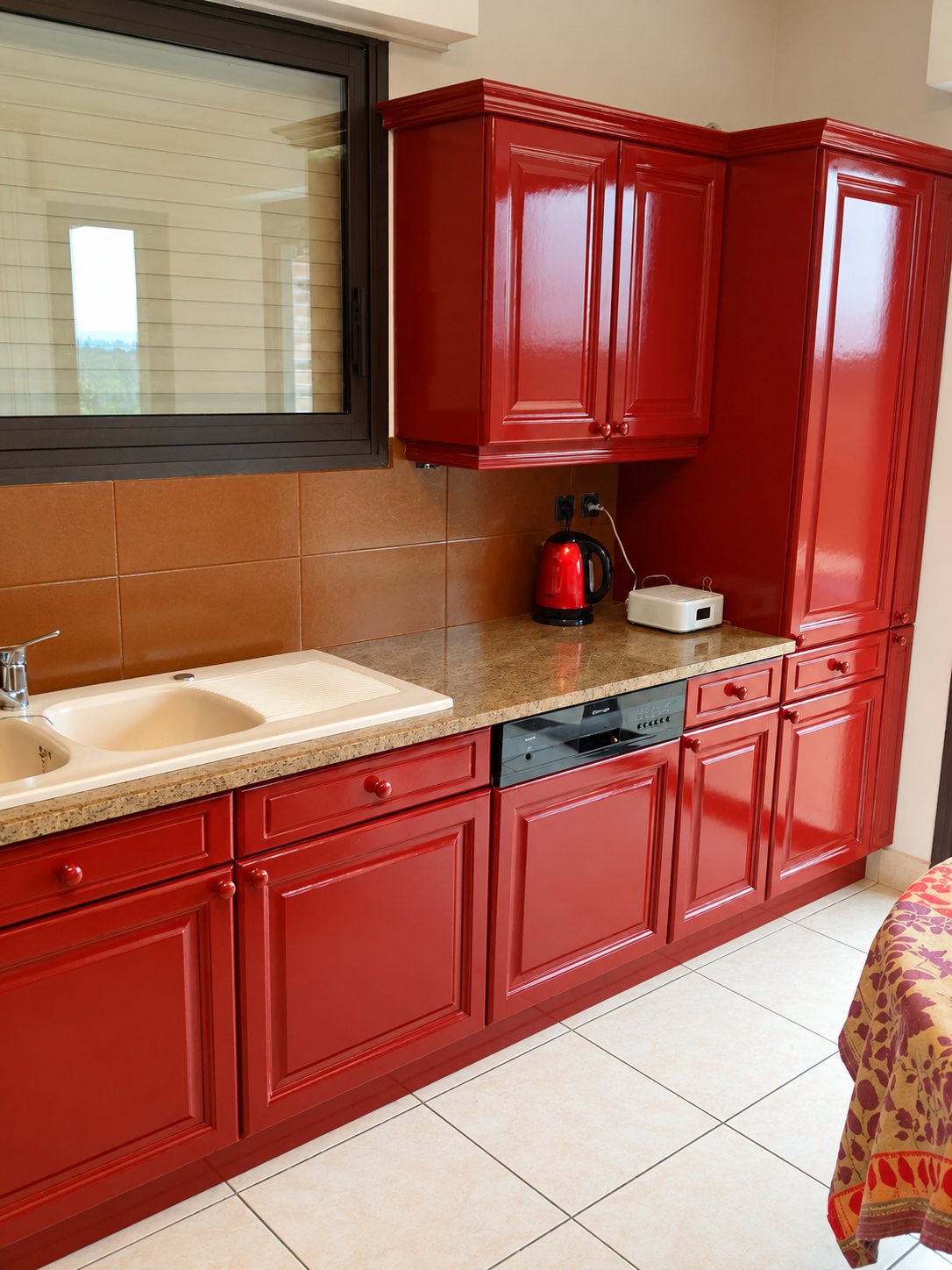 Cuisine équipée aux façades rouges laquées brillantes, avec évier beige, plan de travail en granit, bouilloire rouge et carrelage clair au sol.