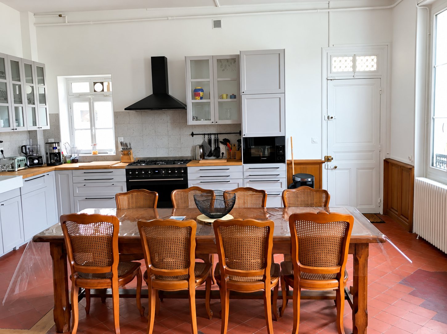 Grande cuisine lumineuse aux placards gris clair, avec piano de cuisson noir hotte noire et longue table en bois entourée de chaises cannées sur un sol en tomettes.
