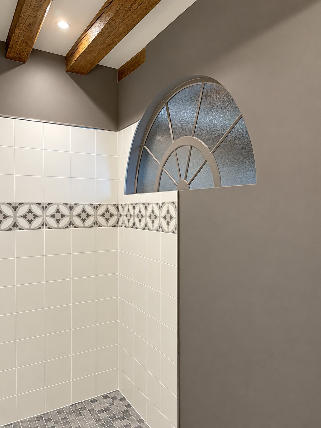 Douche rénovée aux murs carrelés de blanc avec une frise à motifs gris, un sol en mosaïque, des poutres apparentes au plafond et une fenêtre en demi-lune.