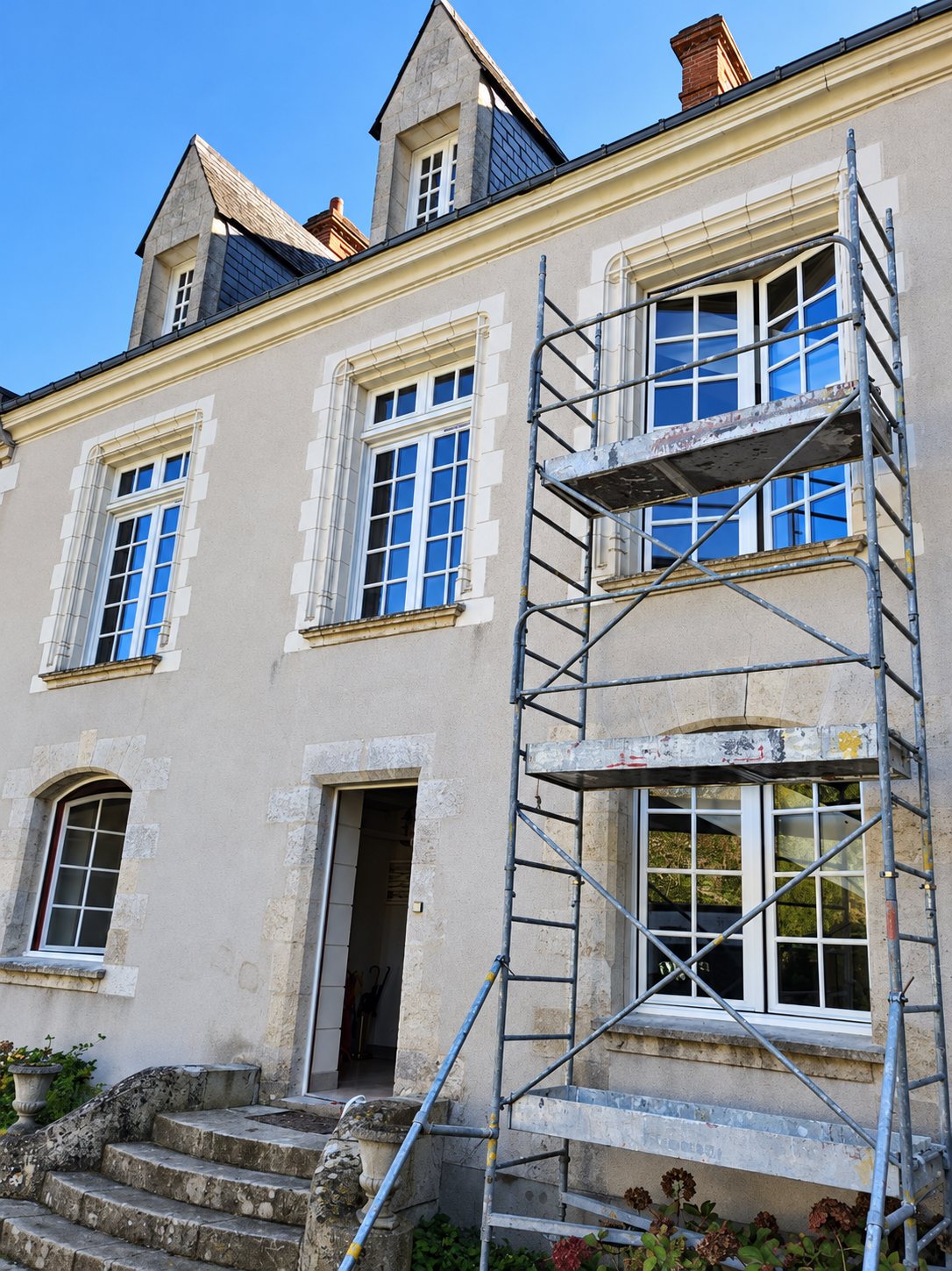 Un échafaudage est dressé devant la façade en pierre claire d'une maison ancienne dont les fenêtres à petits carreaux et les lucarnes sont en cours de rénovation.