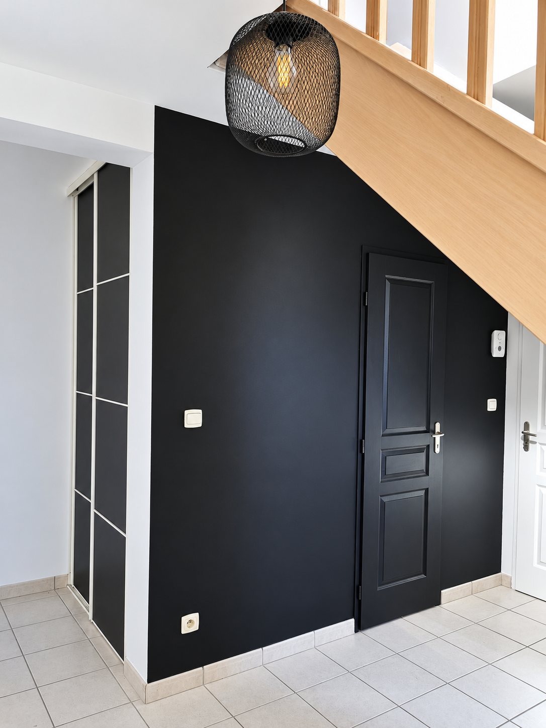 Un mur et une porte peints en noir mat sous un escalier en bois clair, avec un placard à portes coulissantes et une suspension en métal noir.