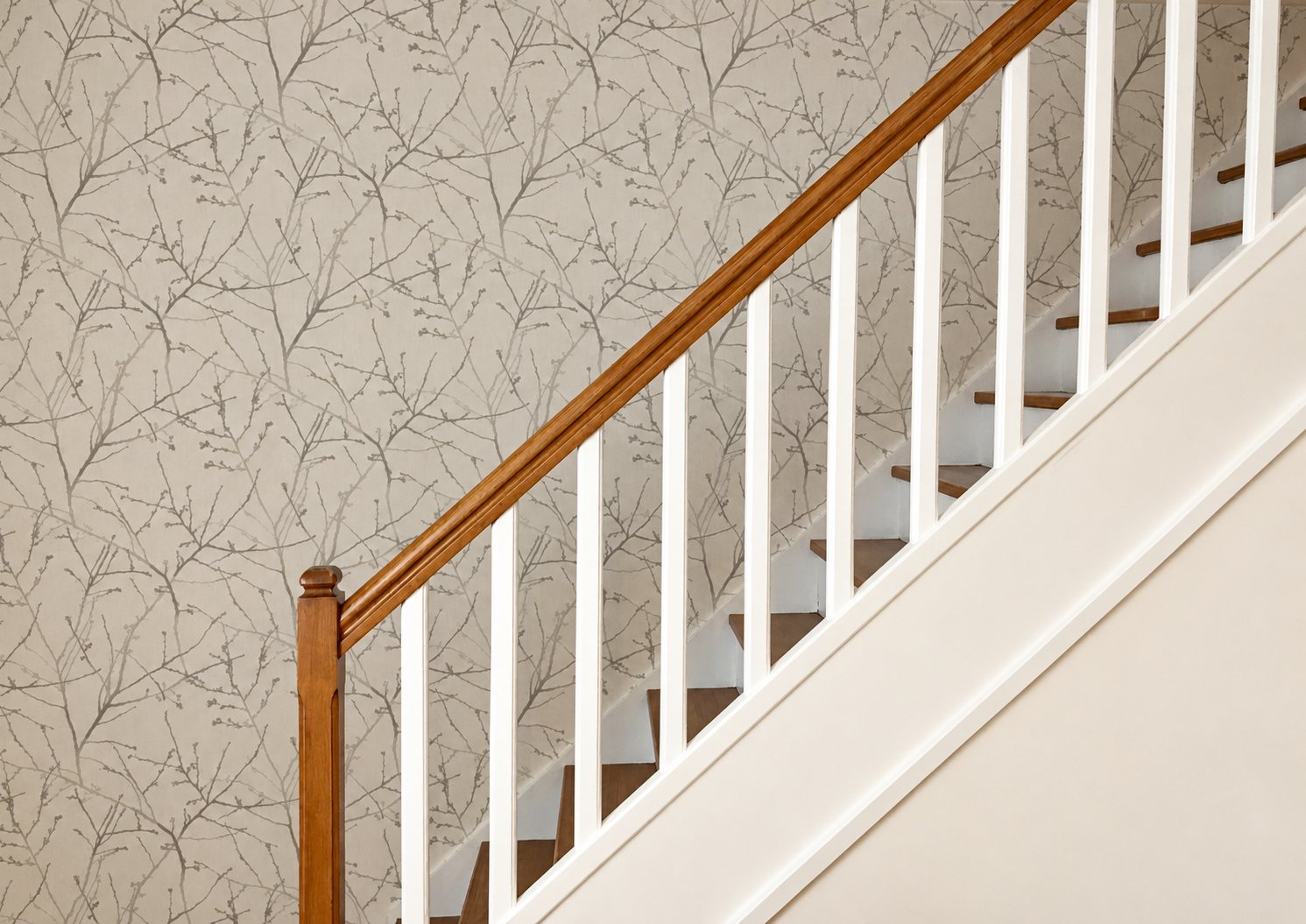 Un escalier aux balustres blancs et à la main courante en bois verni longe un mur tapissé de papier peint gris clair à motif de branches.