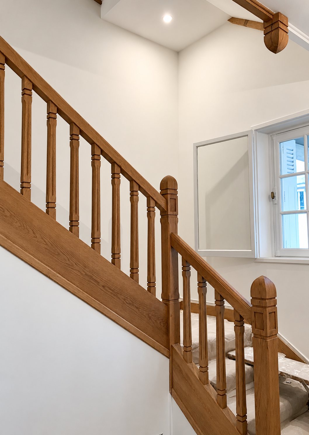 Un escalier en chêne avec rampe et balustres tournés, dans une cage d'escalier aux murs blancs fraîchement repeints, près d'une fenêtre.