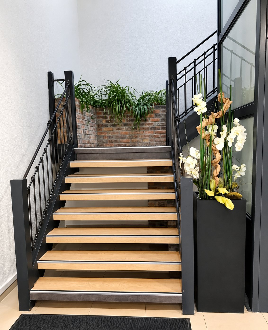 Un escalier intérieur aux marches en bois clair et à la rampe métallique noire, décoré d'un grand vase d'orchidées blanches et de plantes vertes devant un mur de briques.