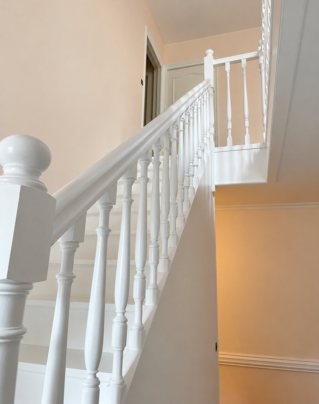 Un escalier intérieur aux balustres tournés et à la rampe peints en blanc, entouré de murs aux teintes rose pâle et pêche.