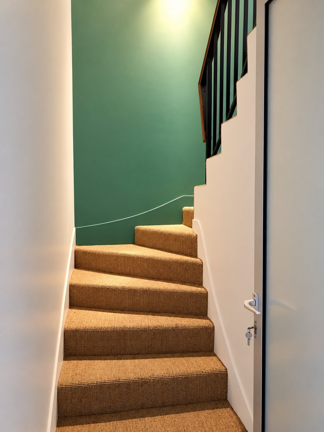 Un escalier étroit et tournant recouvert de moquette en sisal, bordé de murs blancs et d'un mur vert, avec une rampe métallique noire et une porte à droite.
