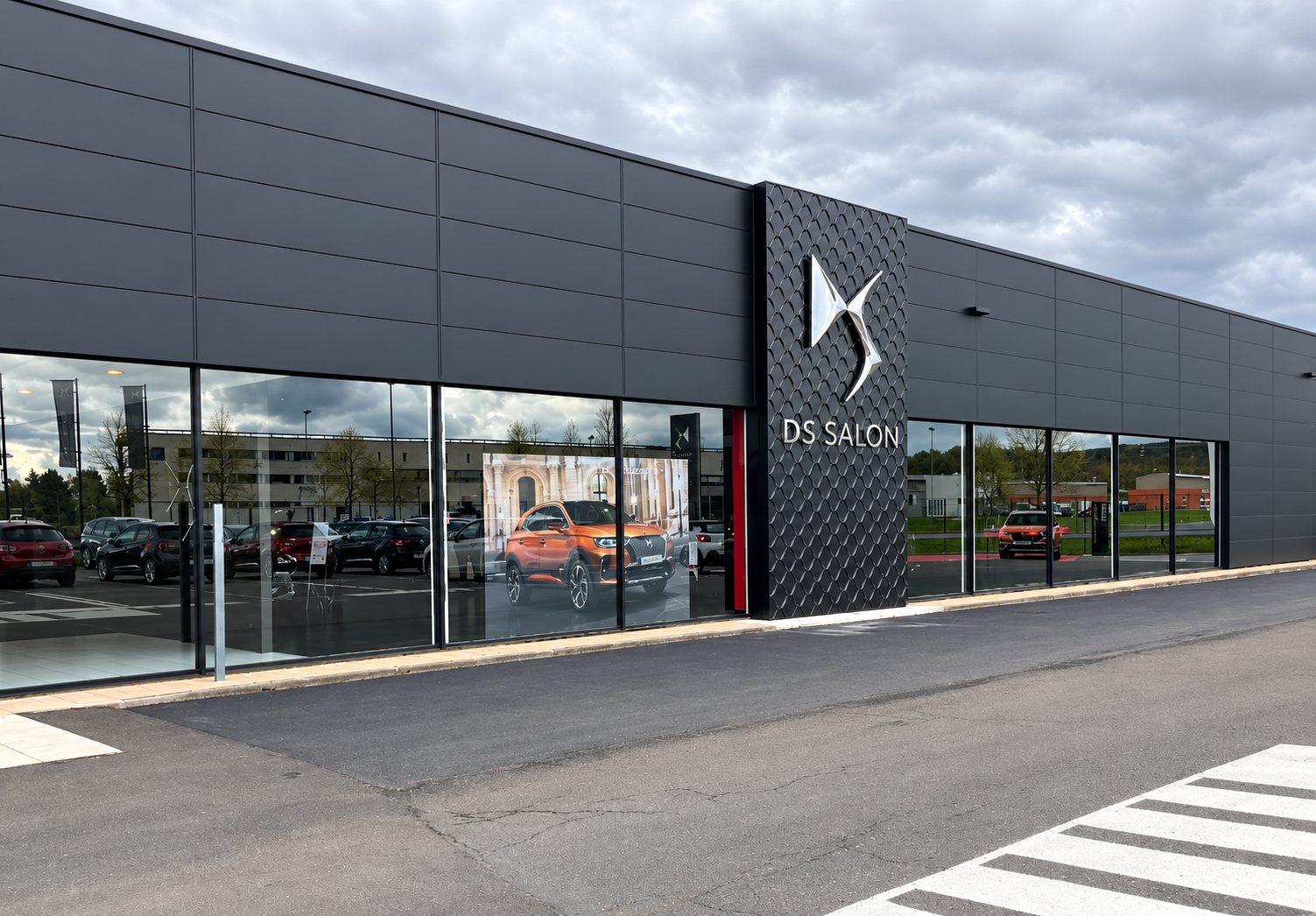 Façade grise anthracite d'une concession DS Salon avec son logo sur un panneau texturé et de grandes baies vitrées donnant sur un parking.