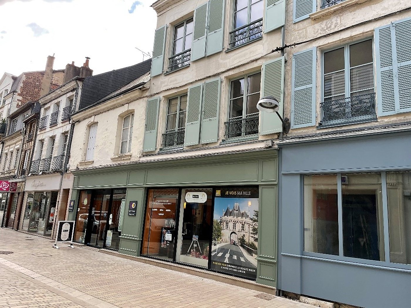 Façade verte d'une boutique avec vitrine affichant une photo de monument, dans une rue pavée bordée d'immeubles anciens aux volets bleu-vert.