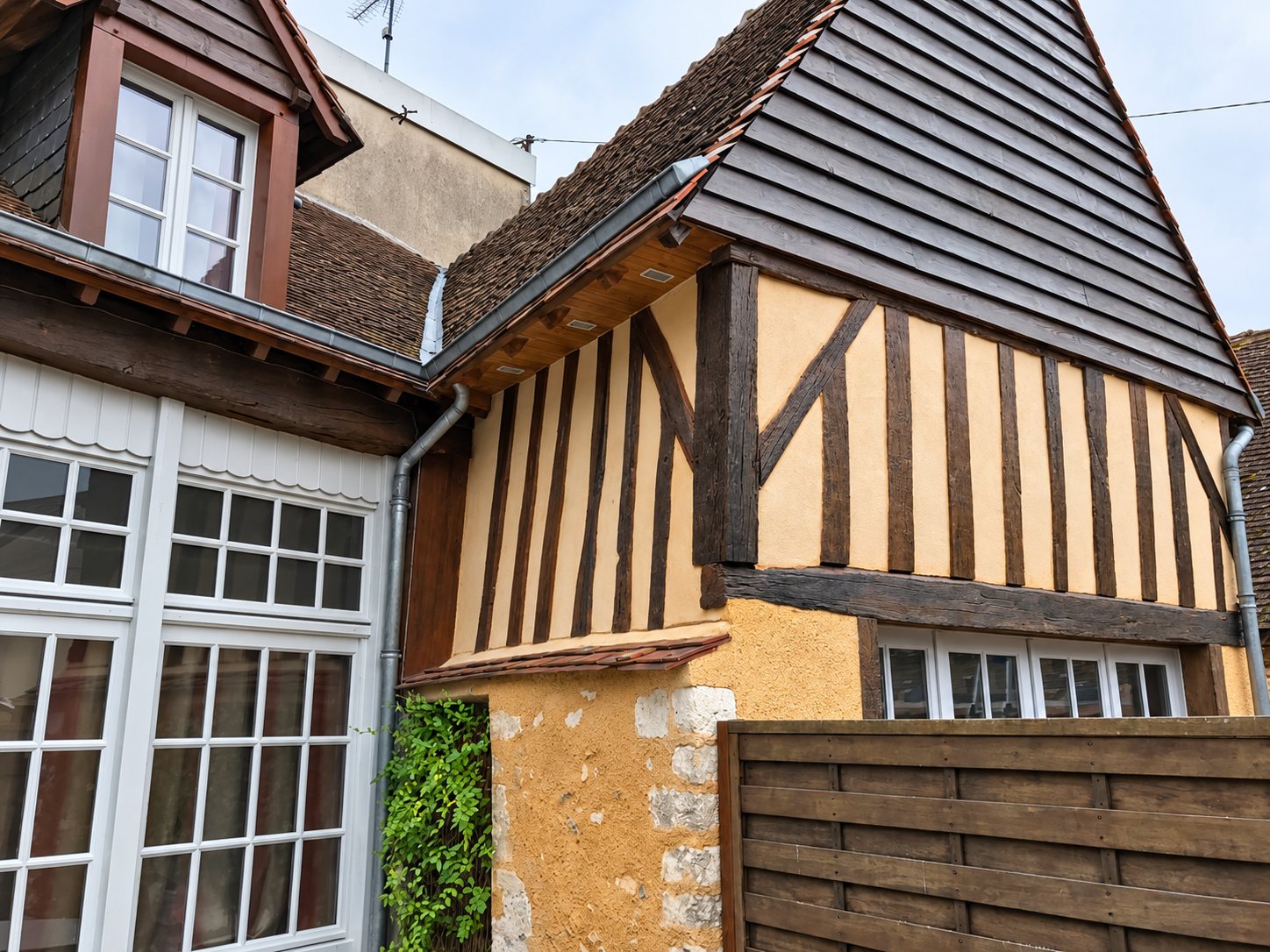 Façade d'une maison ancienne à colombages avec enduit ocre, pignon en bardage de bois sombre, fenêtres blanches à petits carreaux et gouttière en zinc.