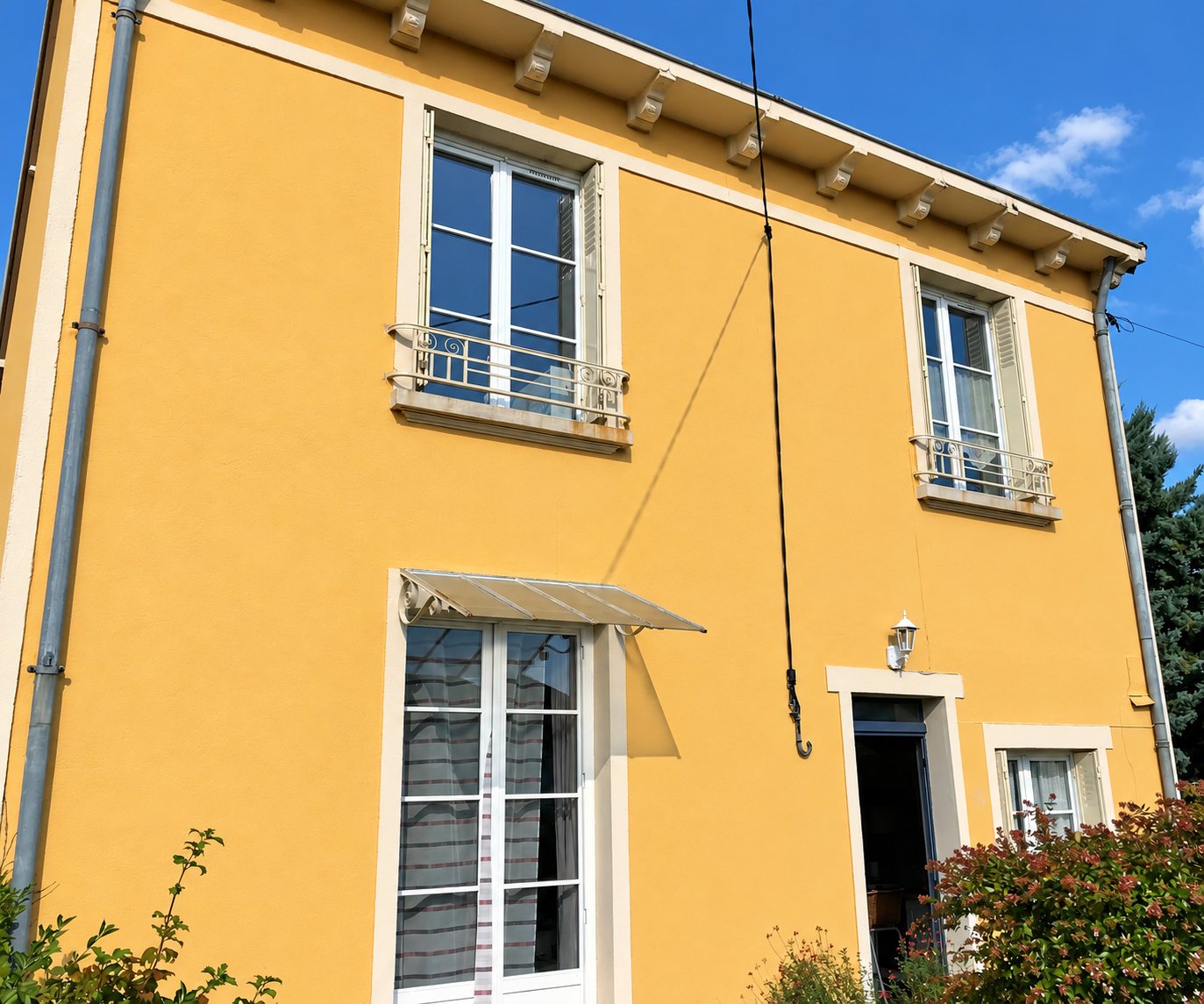 Façade d'une maison enduite en jaune ocre avec encadrements de fenêtres clairs, balcons en fer forgé et petit auvent au-dessus d'une porte-fenêtre.