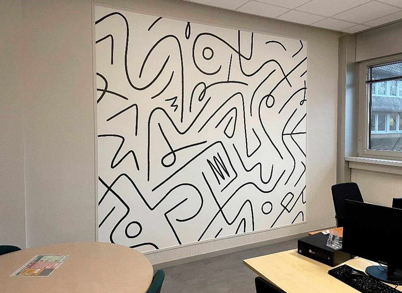 Une grande fresque murale abstraite en lignes noires sur fond blanc décore un bureau équipé d'une table ronde, d'un ordinateur et de chaises.