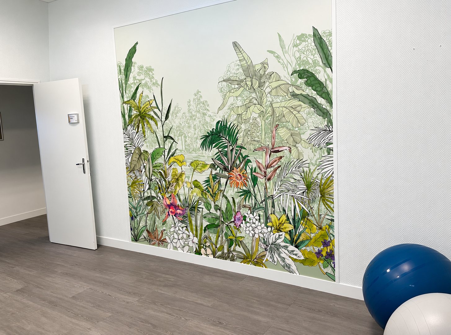 Une grande fresque murale de feuillage tropical vert et de fleurs colorées couvre un mur blanc, à côté d'une porte ouverte et de ballons de gymnastique bleu et gris au sol.
