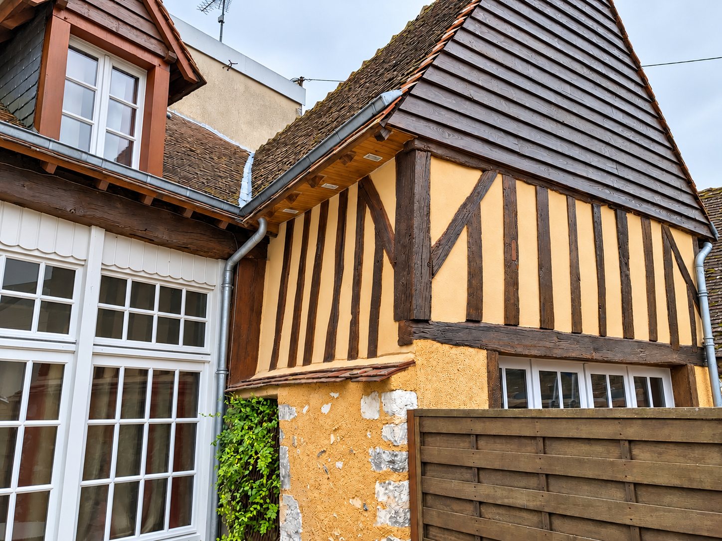 Maison à colombages rénovée : enduit ocre, pignon en bardage de bois sombre, fenêtres blanches à petits carreaux et gouttière en zinc.