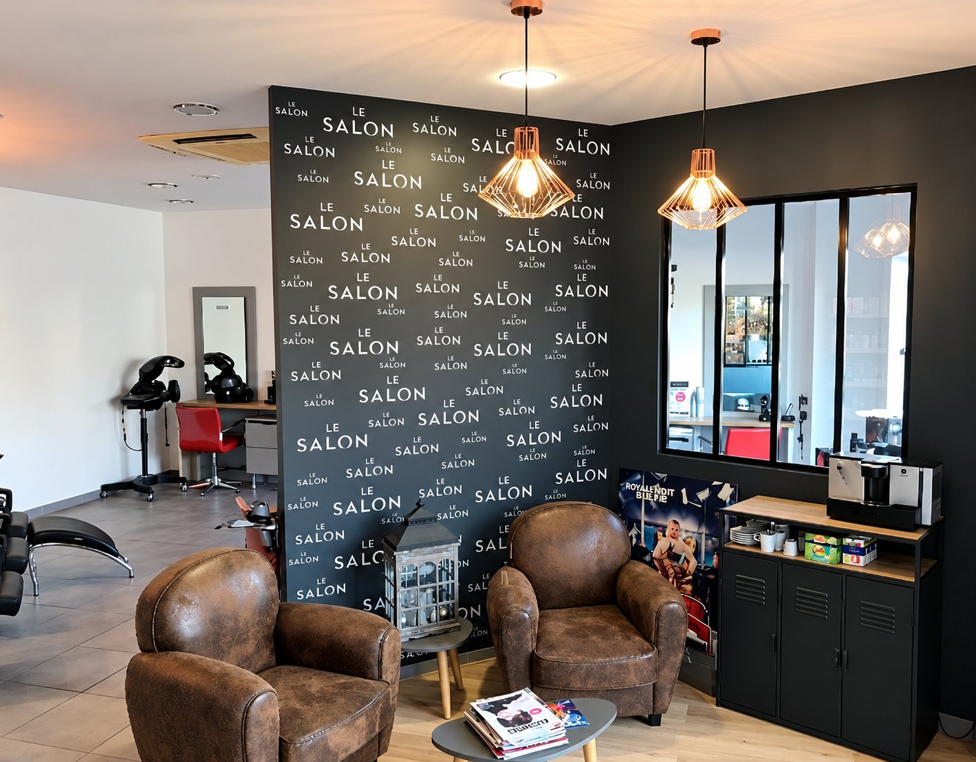 Espace d'accueil d'un salon de coiffure avec deux fauteuils en cuir marron, un mur noir imprimé du nom du salon et des suspensions cuivrées.