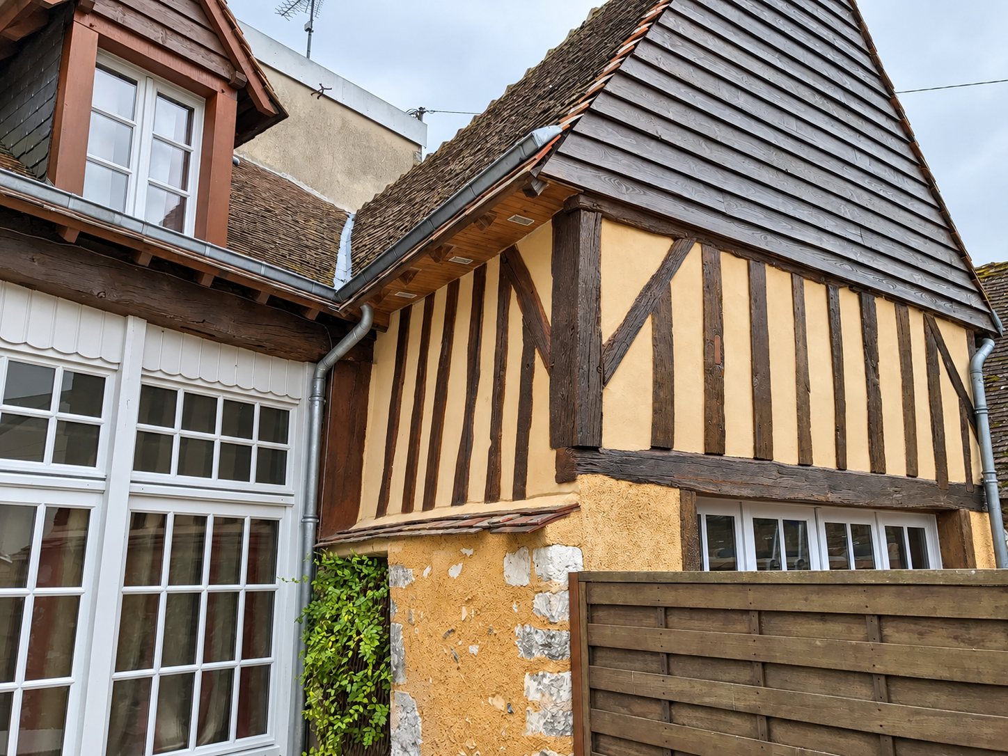 Façade d'une maison ancienne à colombages avec enduit ocre, pignon en bardage bois sombre, gouttières en zinc et grandes fenêtres blanches.