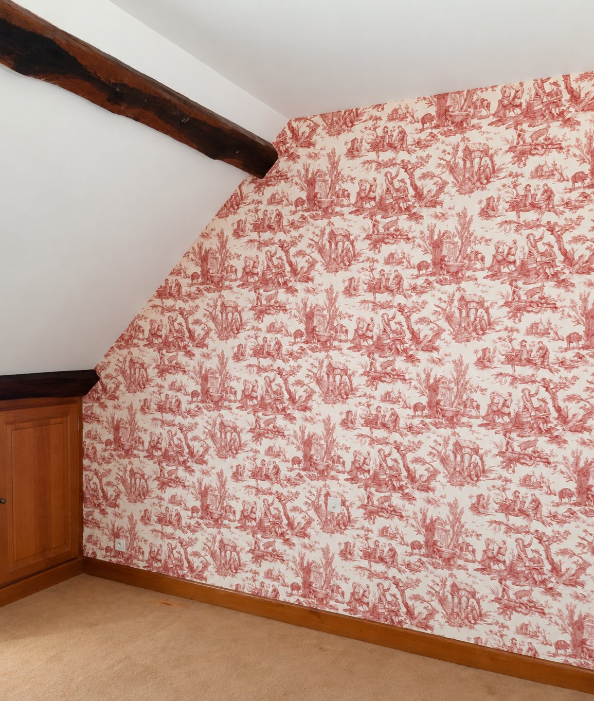 Pièce sous les combles dont le mur incliné est recouvert d'un papier peint toile de Jouy rouge sur fond crème, avec une poutre en bois et un placard.
