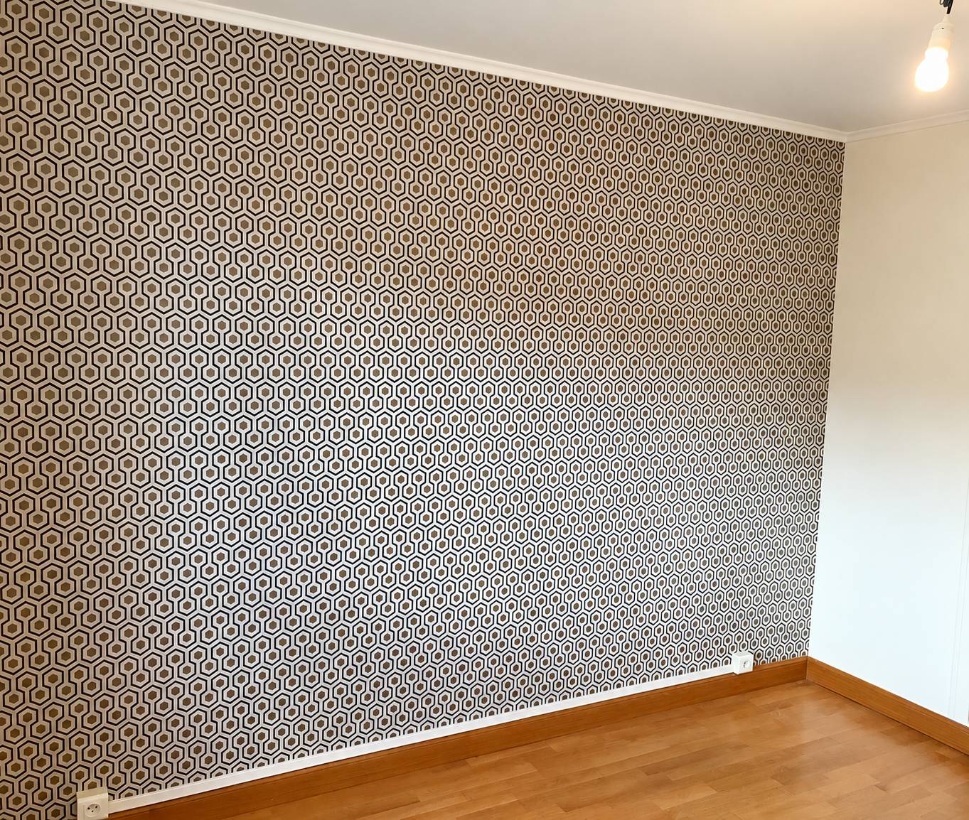 Une pièce vide au parquet en bois dont un mur est recouvert d'un papier peint à motif hexagonal noir, blanc et beige.