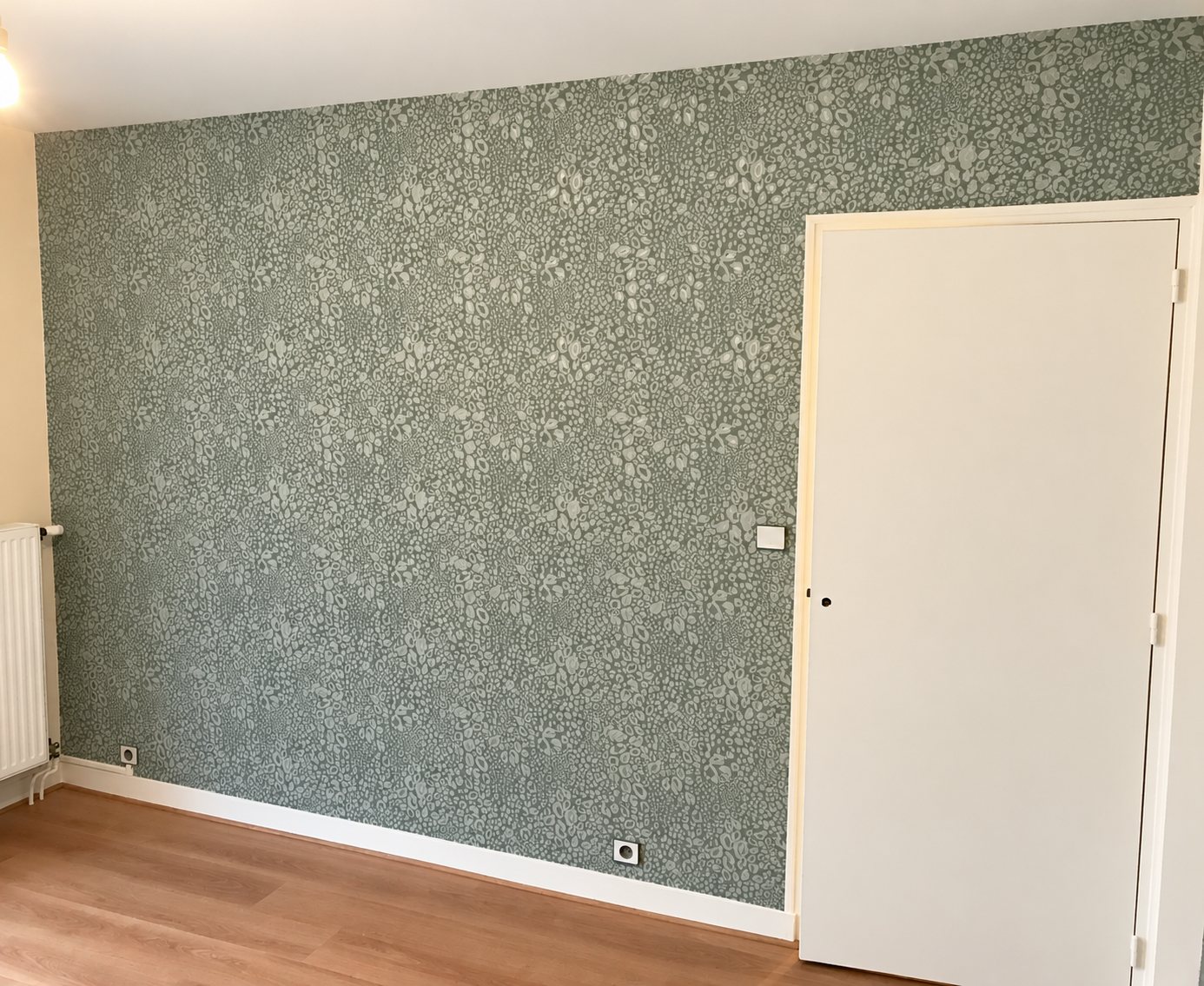 Pièce rénovée avec un mur tapissé de papier peint vert sauge à motif organique, une porte blanche, un radiateur et un parquet en bois.