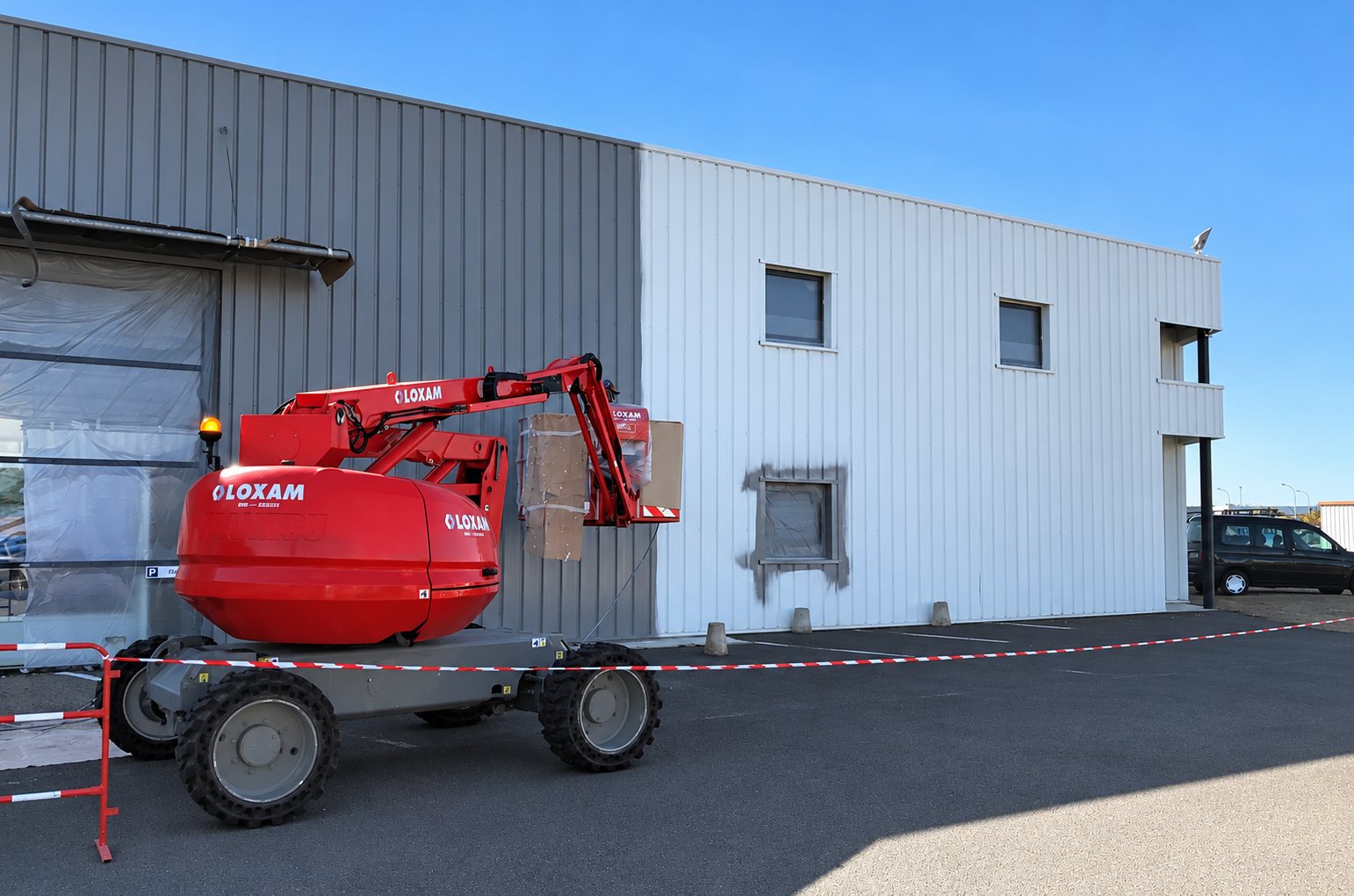 Une nacelle élévatrice rouge Loxam est positionnée devant la façade d'un bâtiment industriel dont la moitié du bardage métallique a déjà été nettoyée, avec un ruban de balisage au sol.