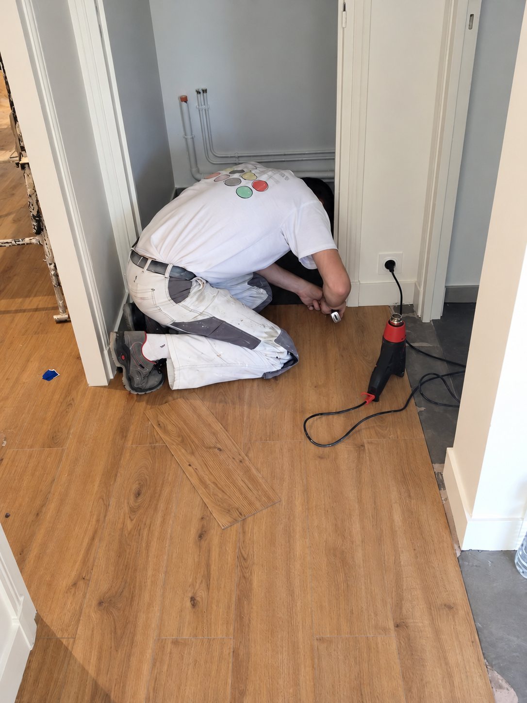 Un ouvrier en tenue blanche est agenouillé et pose un revêtement de sol imitation parquet dans un petit local, avec un décapeur thermique posé à côté.