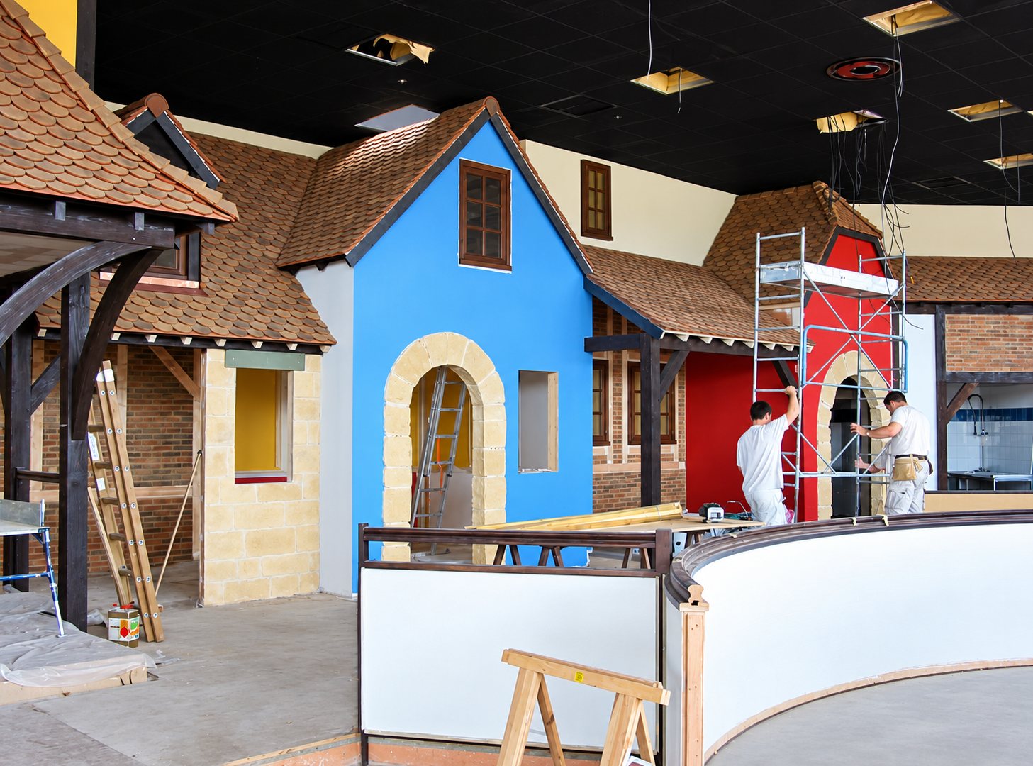Deux ouvriers travaillent sur un chantier intérieur où des façades de maisons bleue et rouge au toit de tuiles forment un décor de village.