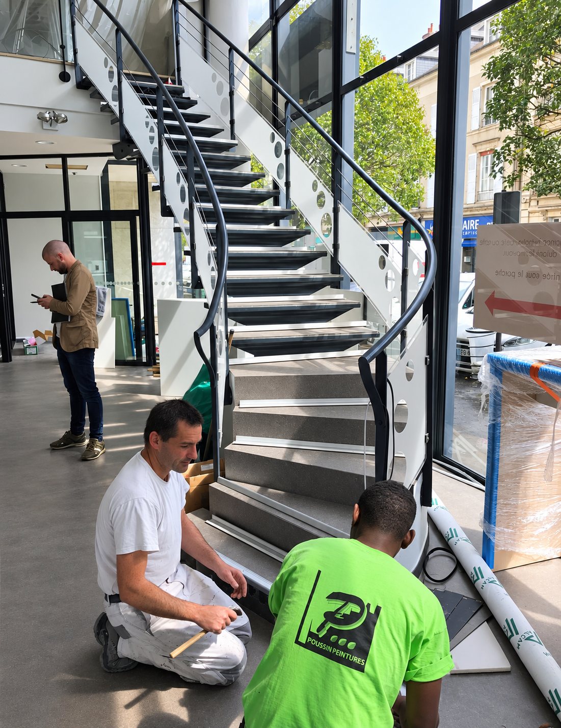 Deux ouvriers agenouillés travaillent au pied d'un escalier courbe à garde-corps métallique et verre, tandis qu'un homme consulte son téléphone dans un hall vitré.