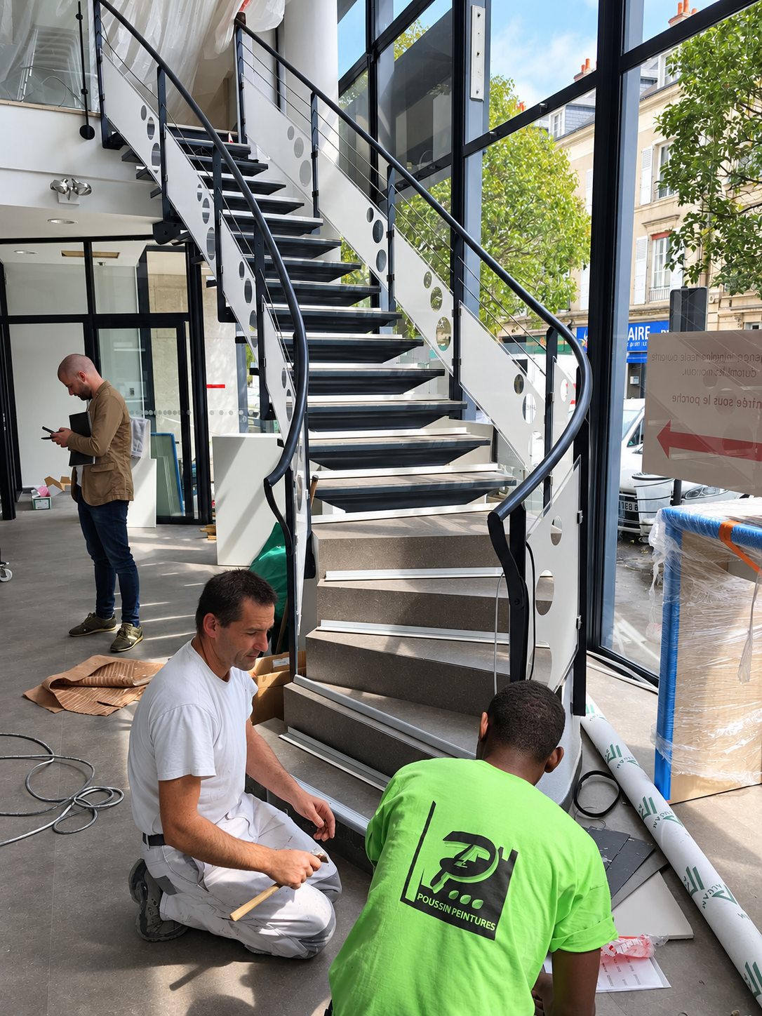 Deux ouvriers accroupis travaillent au pied d'un escalier courbe à garde-corps métallique, tandis qu'un homme consulte des documents à l'arrière-plan dans un local aux grandes baies vitrées.