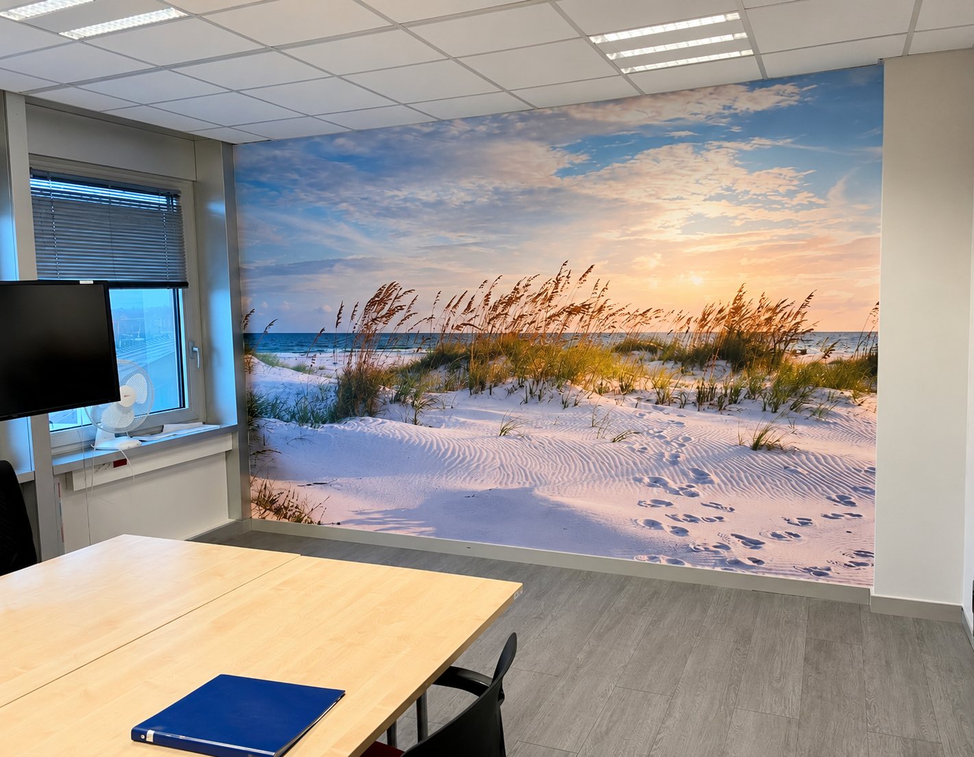 Une salle de réunion de bureau dont un mur est recouvert d'une grande photo de dunes de sable, d'herbes et de mer au coucher du soleil, avec une table en bois au premier plan.