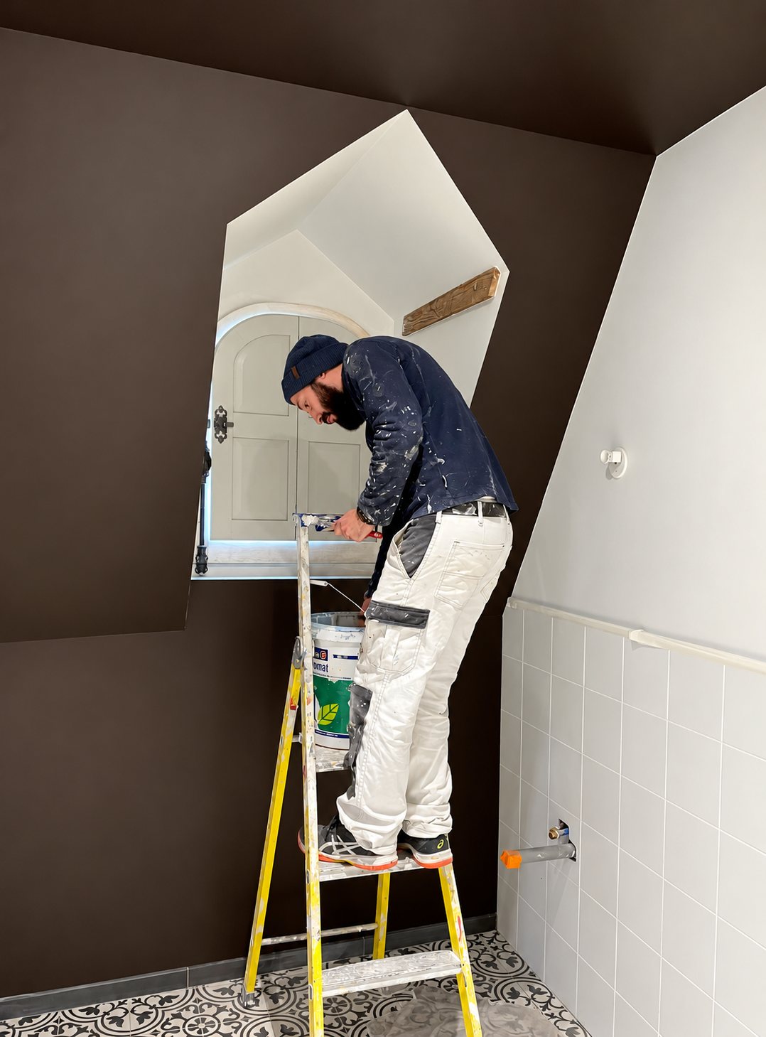 Un peintre en tenue tachée de blanc se tient sur un escabeau jaune et peint les murs bruns d'une salle de bain sous les combles, près d'une lucarne.