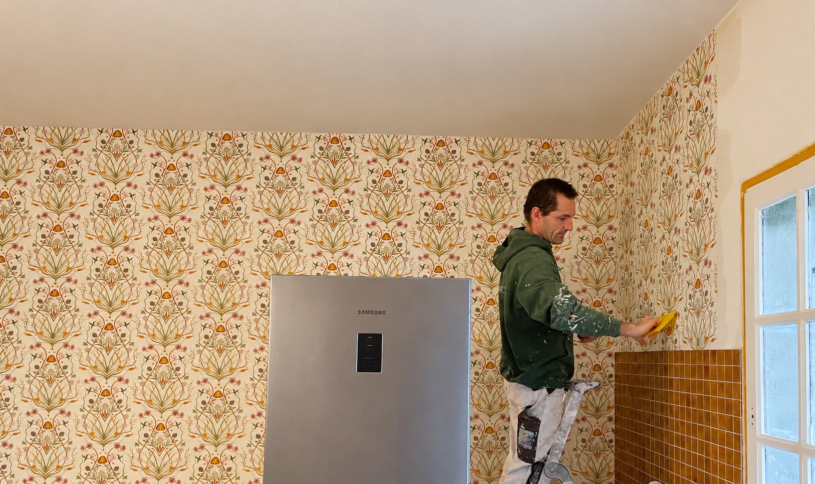 Un peintre en sweat vert lisse un papier peint à motifs floraux crème dans une cuisine, à côté d'un réfrigérateur Samsung et d'une fenêtre.