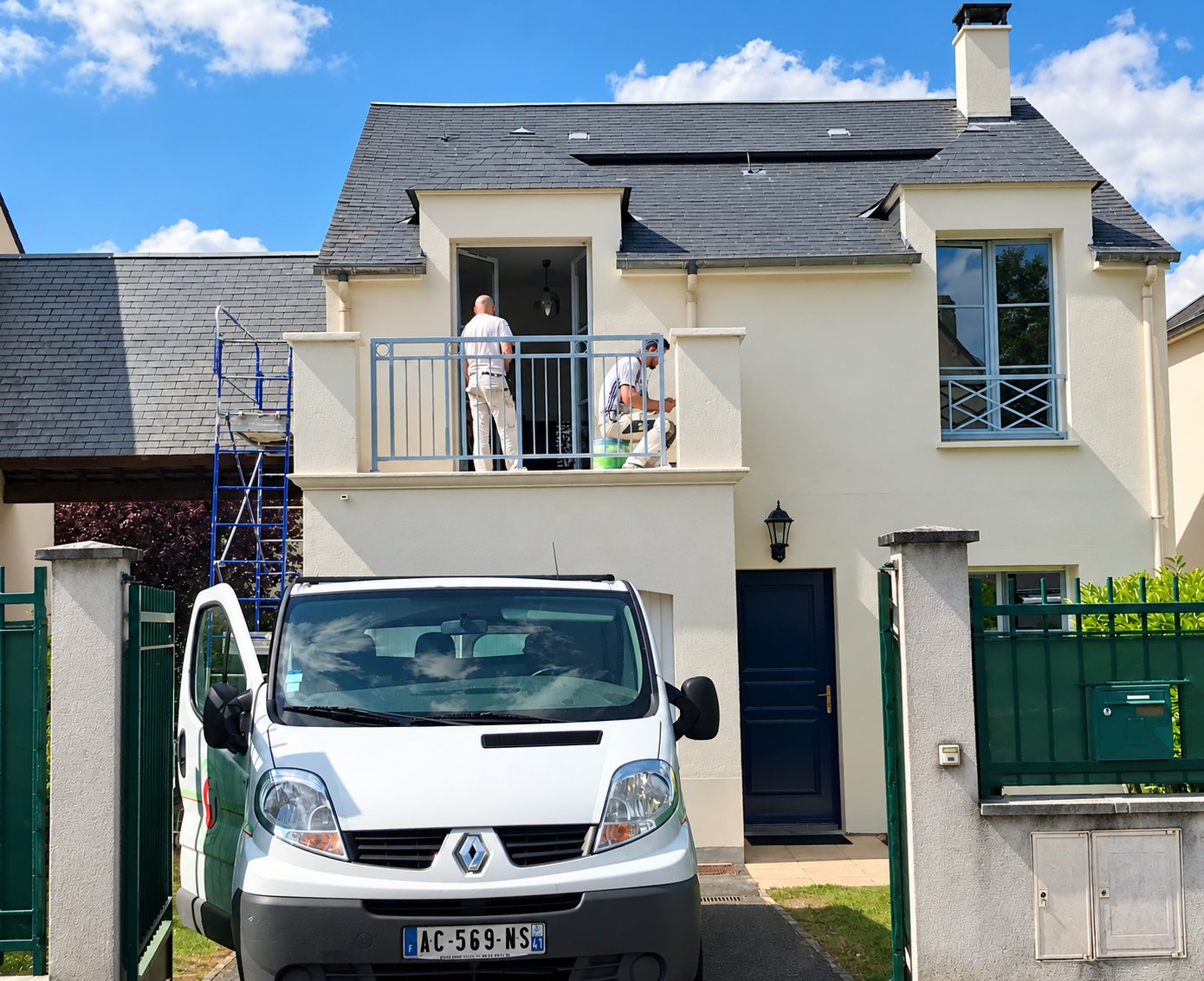 Deux peintres en tenue blanche travaillent sur le balcon d'une maison à façade crème, avec un échafaudage bleu et une camionnette blanche garée devant le portail.