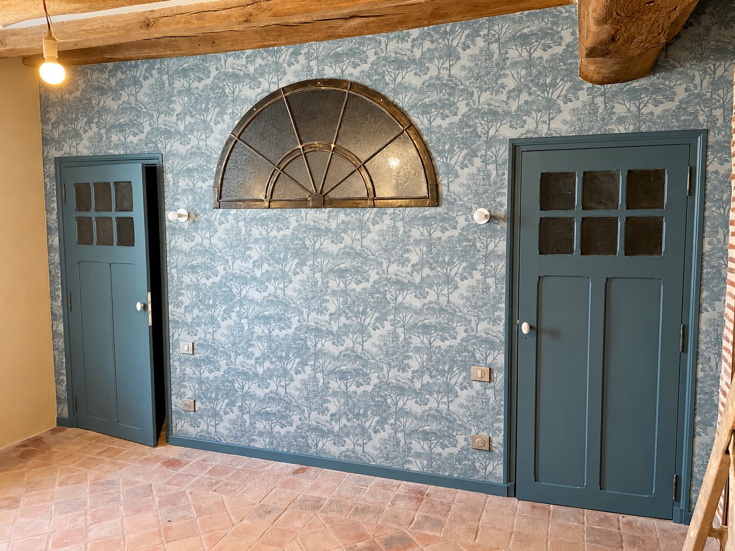 Pièce rénovée avec un papier peint motif arbres gris, deux portes vert foncé vitrées, une imposte en demi-lune et un sol en tomettes.