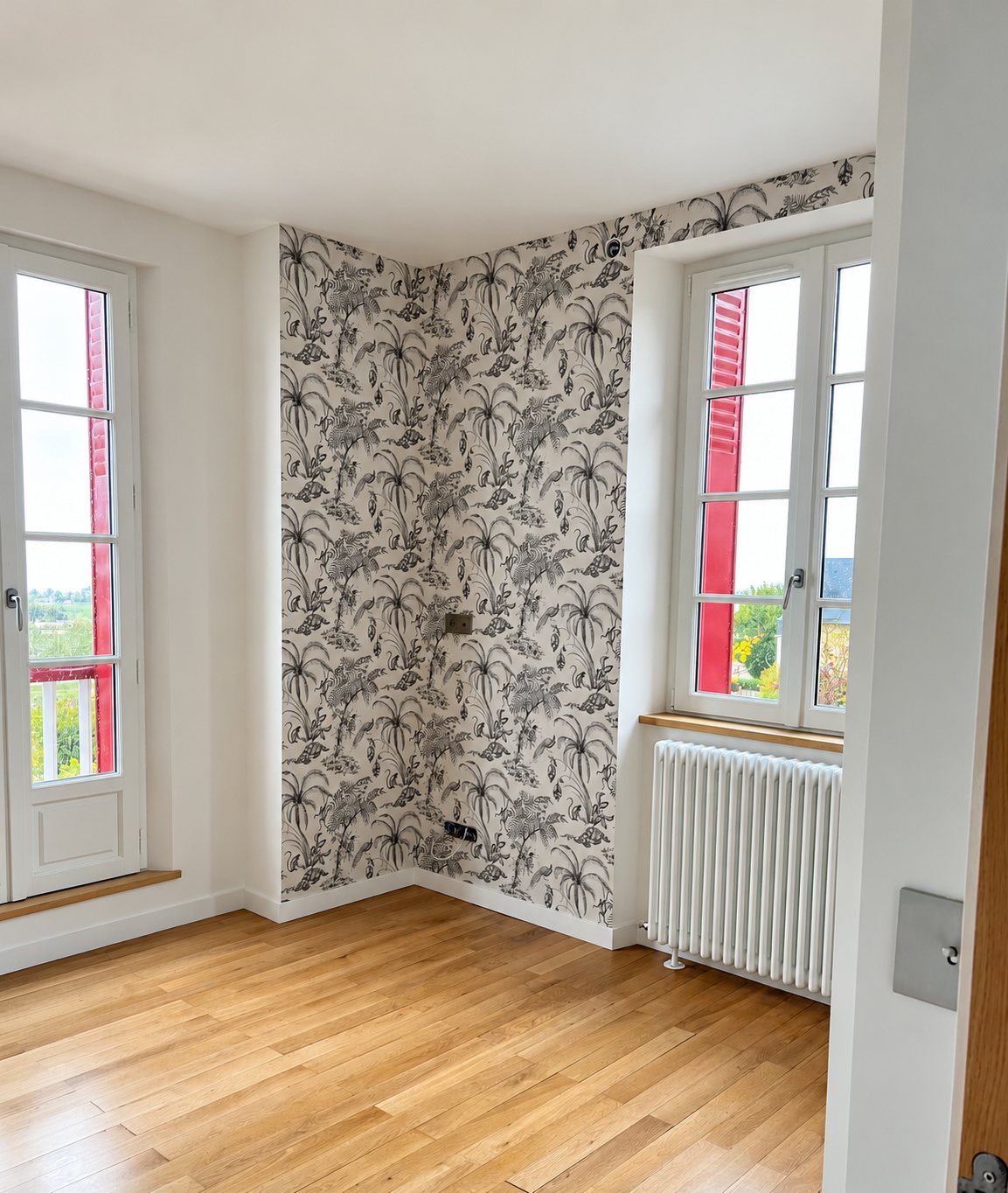 Pièce rénovée avec un papier peint à motifs de plantes tropicales noir et blanc, un parquet en chêne clair, un radiateur blanc et deux fenêtres à volets rouges.