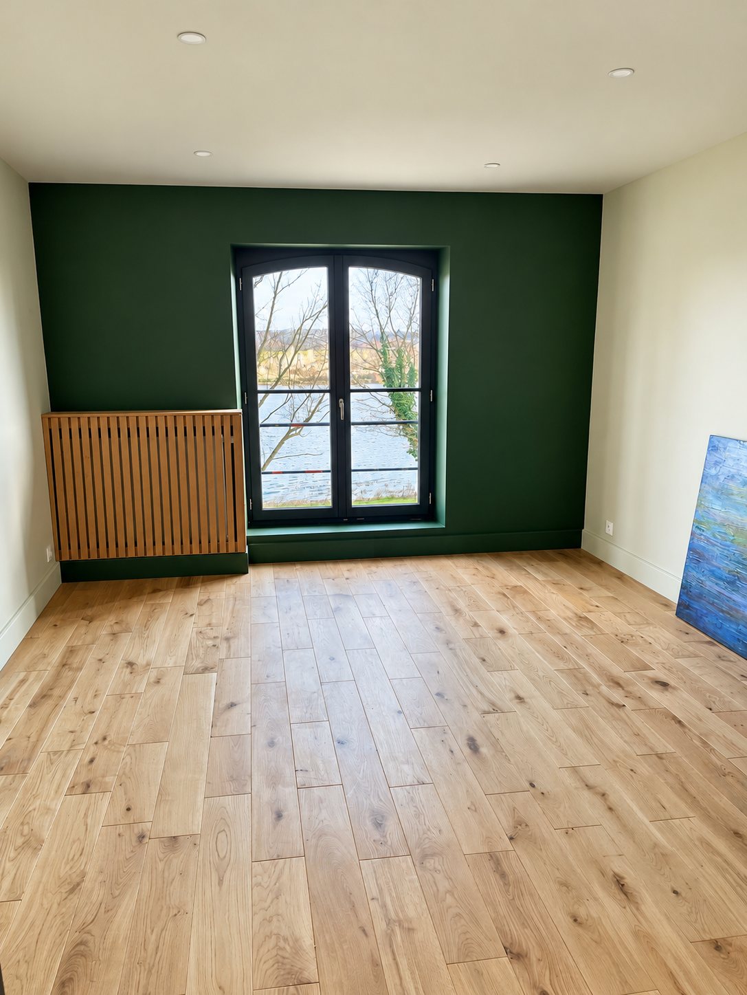 Pièce vide rénovée avec un parquet en chêne clair, un mur vert foncé, une porte-fenêtre noire et un cache-radiateur en bois à lattes.