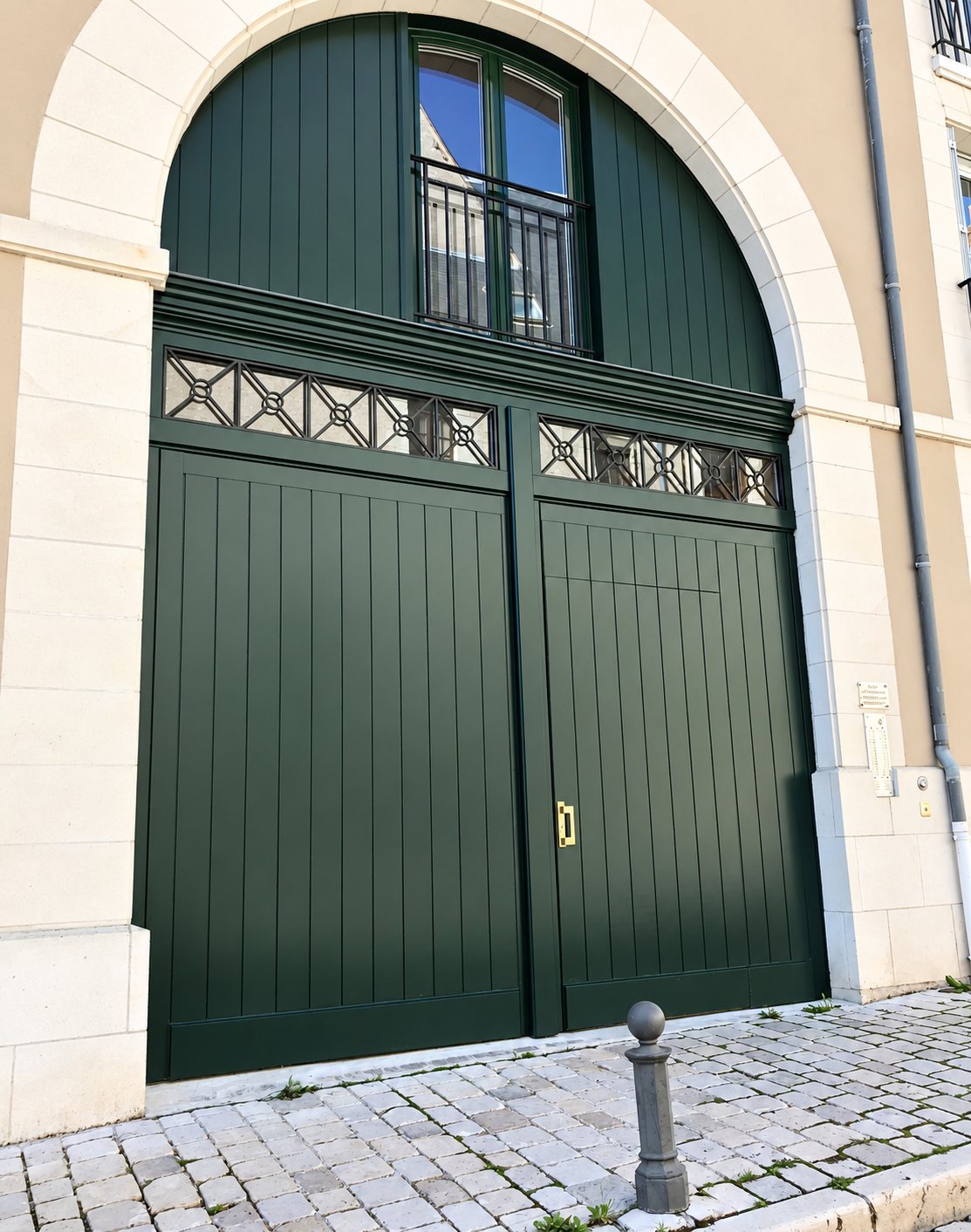 Une grande porte cochère en bois vert foncé à deux battants, surmontée d'une imposte ajourée et d'une fenêtre, sous une arche en pierre claire au-dessus d'un trottoir pavé.