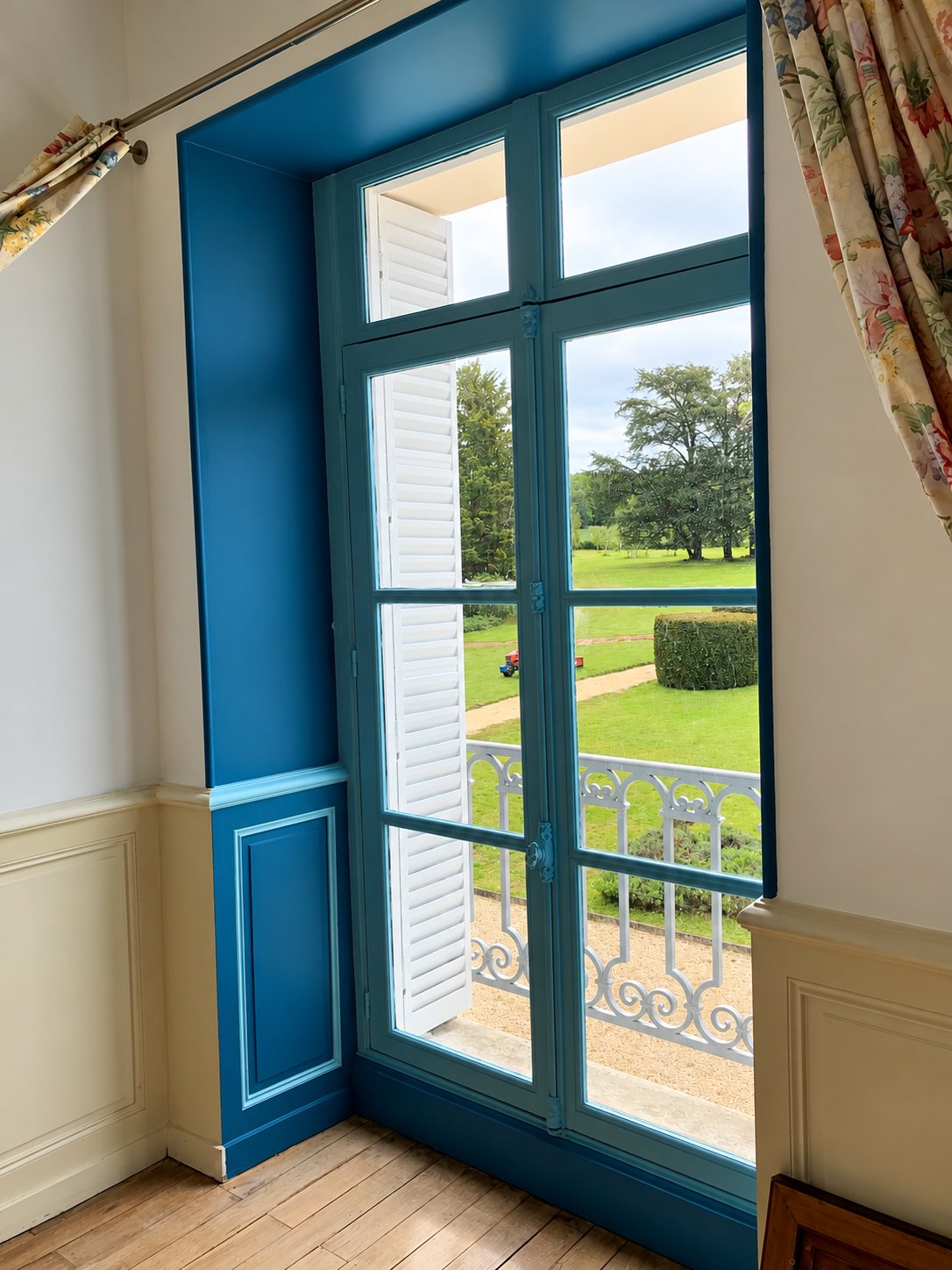 Une porte-fenêtre à petits carreaux et son embrasure peintes en bleu, avec volets blancs ouverts sur un balcon et un parc arboré.