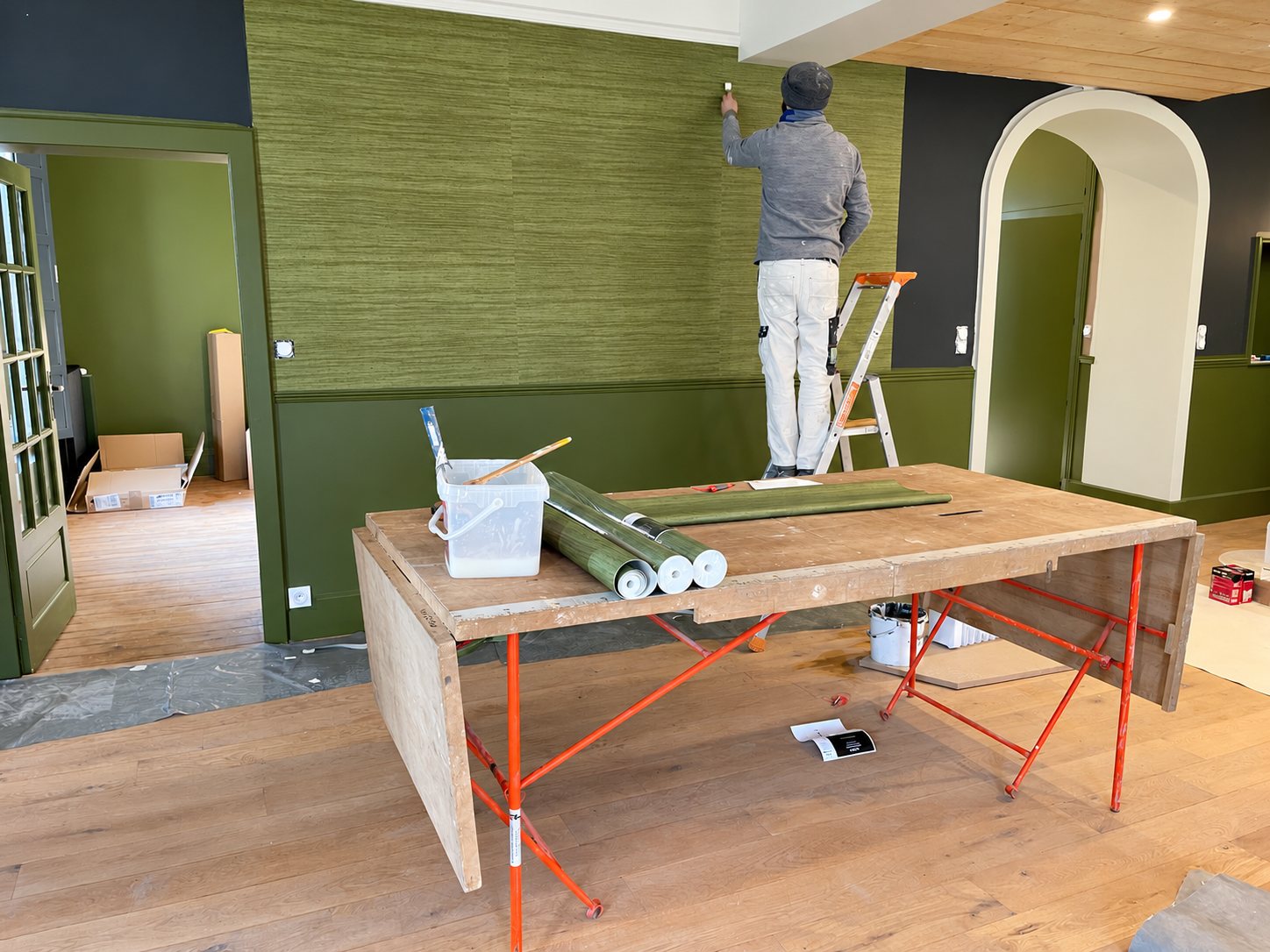 Un artisan pose du papier peint vert texturé sur un mur d'une grande pièce en rénovation, avec une table de collage et des rouleaux au premier plan.