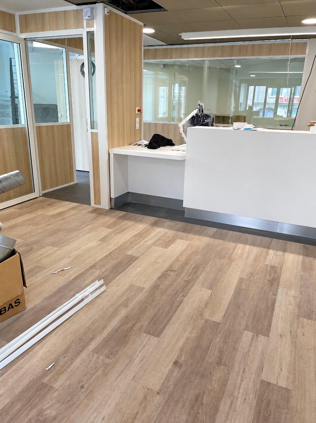 Intérieur en cours de rénovation avec un sol imitation bois clair, des cloisons vitrées aux panneaux de bois et un comptoir d'accueil blanc.