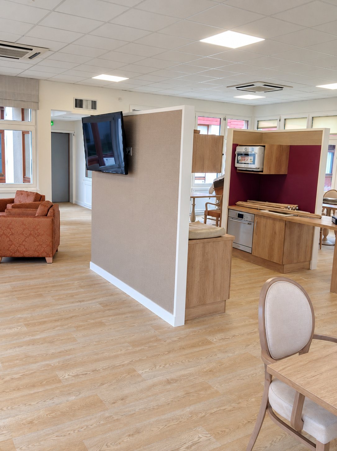 Grande salle lumineuse au sol clair avec cloison beige portant un téléviseur, salon aux fauteuils orangés et coin cuisine en bois et bordeaux.