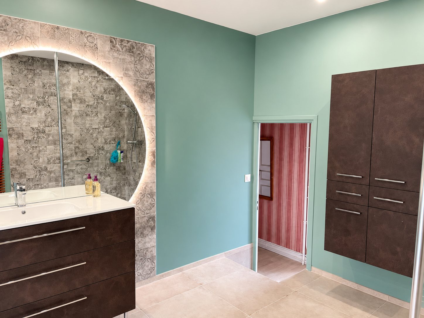 Salle de bain rénovée aux murs vert d'eau, avec un grand miroir rond lumineux, un meuble vasque brun foncé, une colonne de rangement suspendue et un carrelage beige au sol.