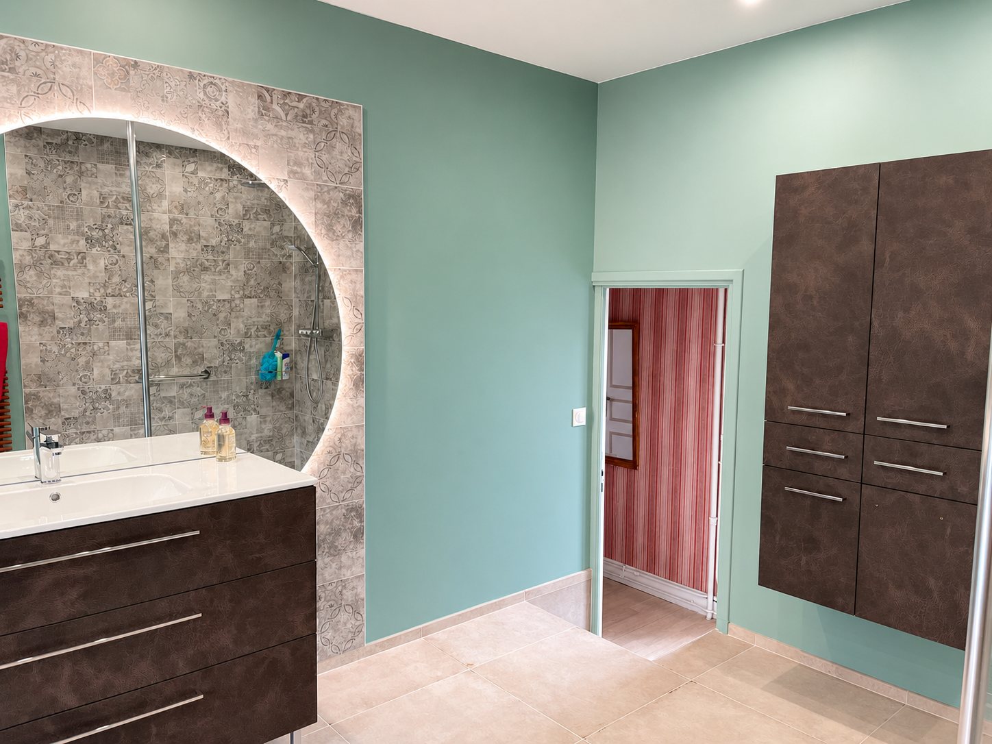Salle de bains rénovée aux murs vert sauge, avec meuble vasque brun, grand miroir rond lumineux devant une douche carrelée et colonne de rangement suspendue.