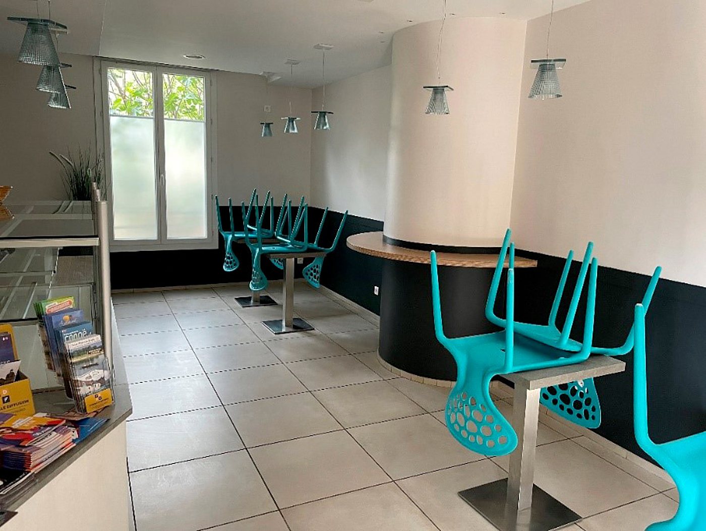 Intérieur d'une petite salle avec des chaises hautes turquoise retournées sur des tables en inox, des suspensions grises, un comptoir vitré avec des brochures et un sol carrelé clair.